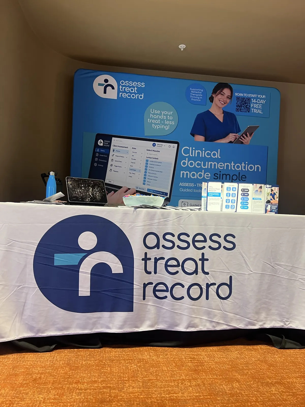 The Assess Treat Record stand at the AMT annual conference: a blue banner reading Clinical documentation made simple above a table with the Assess Treat Record logo, brochures and an iPad